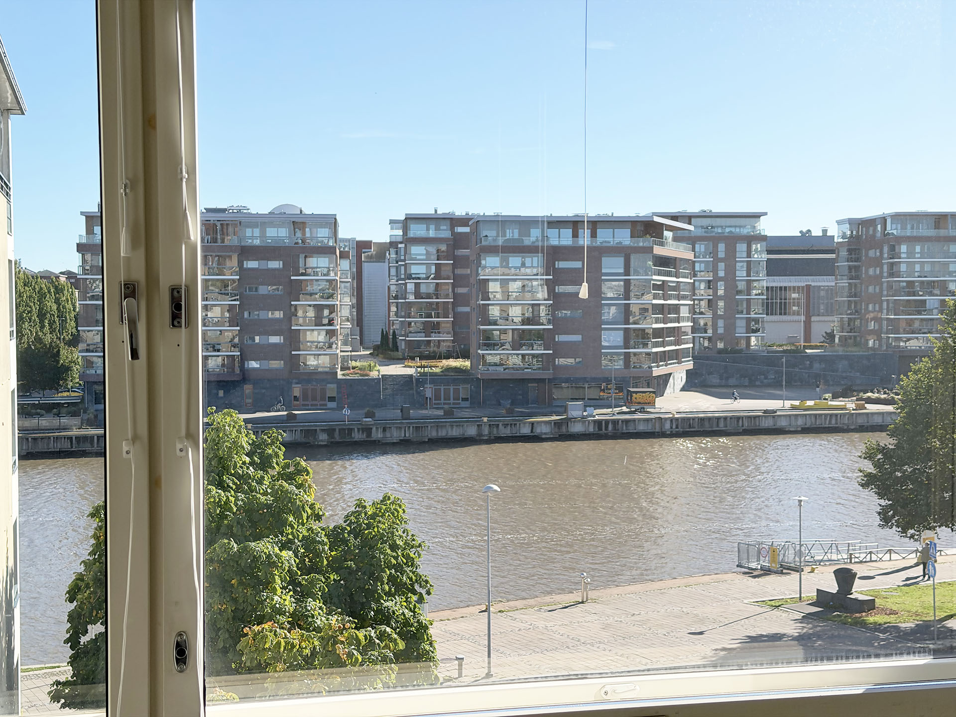 Näkymä ikkunasta joelle, jonka vastarannalla kohoavat suuret, modernit tiilitalot.