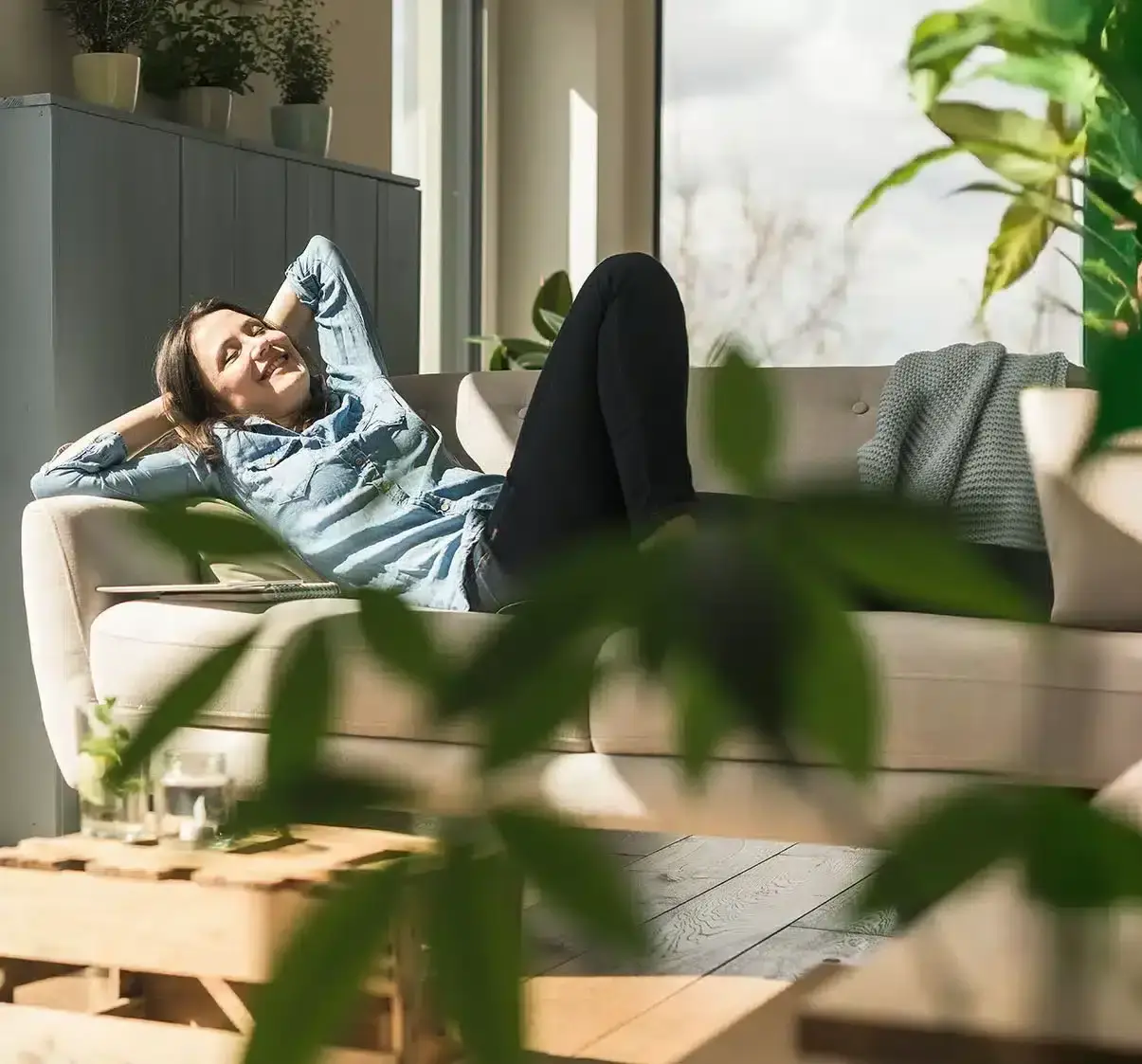 Nainen rentoutuu beigeellä sohvalla kädet pään takana ja hymyilee. Auringonvalo tulvii sisään suurista ikkunoista, ja huonekasvit koristavat kodikasta, modernia olohuonetta.