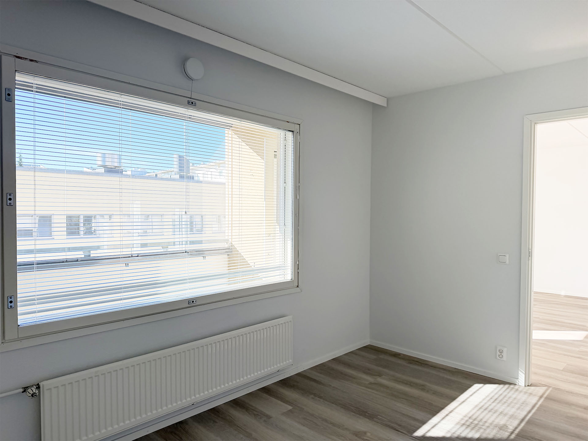 Valoisa, tyhjä huone, jossa on vaalea puunvärinen lattia, suuri ikkuna valkoisilla kaihtimilla sekä avoin oviaukko, joka johtaa toiseen huoneeseen.