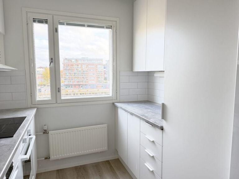 Minimalistinen valkoinen keittiö, jonka ikkunasta avautuu näkymä kerrostaloon; keittiössä on harmaat työtasot, valkoiset kaapit ja ikkunan alla lämpöpatteri.