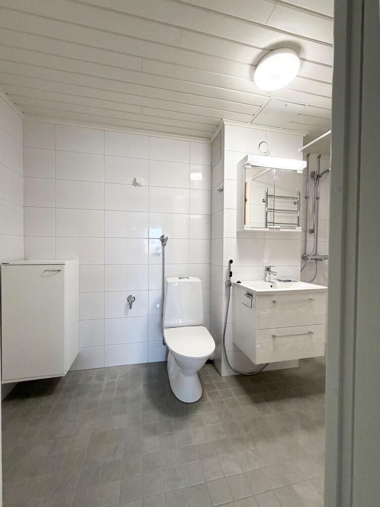 Valoisa kylpyhuone, jossa on valkoisella laatoitetut seinät, seinälle kiinnitetty pesuallas ja WC.