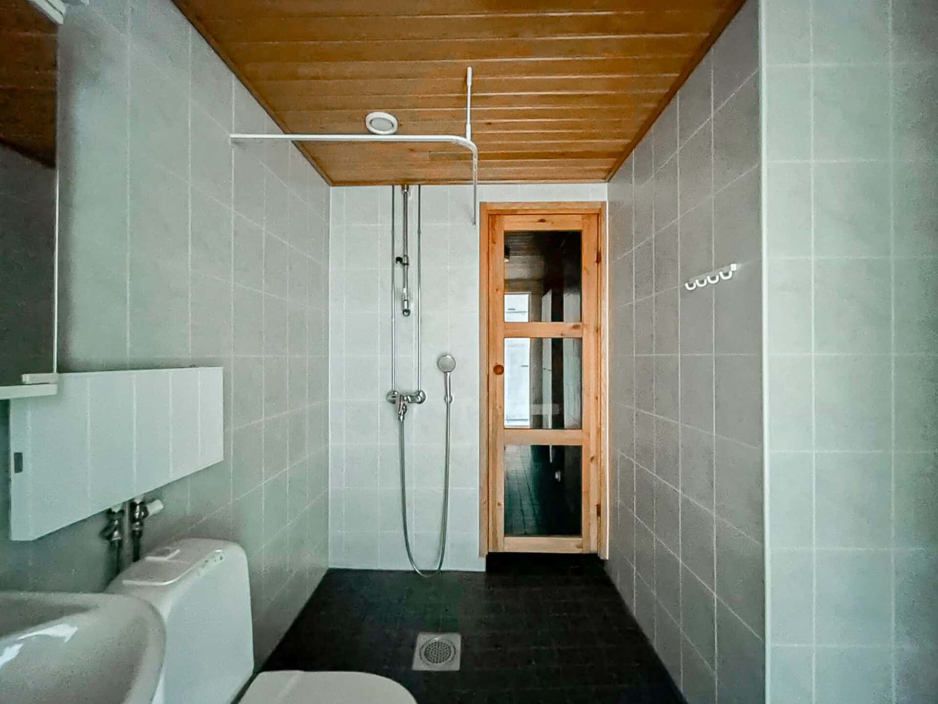 Moderni kylpyhuone, jossa on harmaat laatoitetut seinät, wc, suihkualue käsisuihkupäällä sekä puinen ovi, joka johtaa saunaan. Katto on puupaneloitu, ja oikealla seinällä on koukkuja.