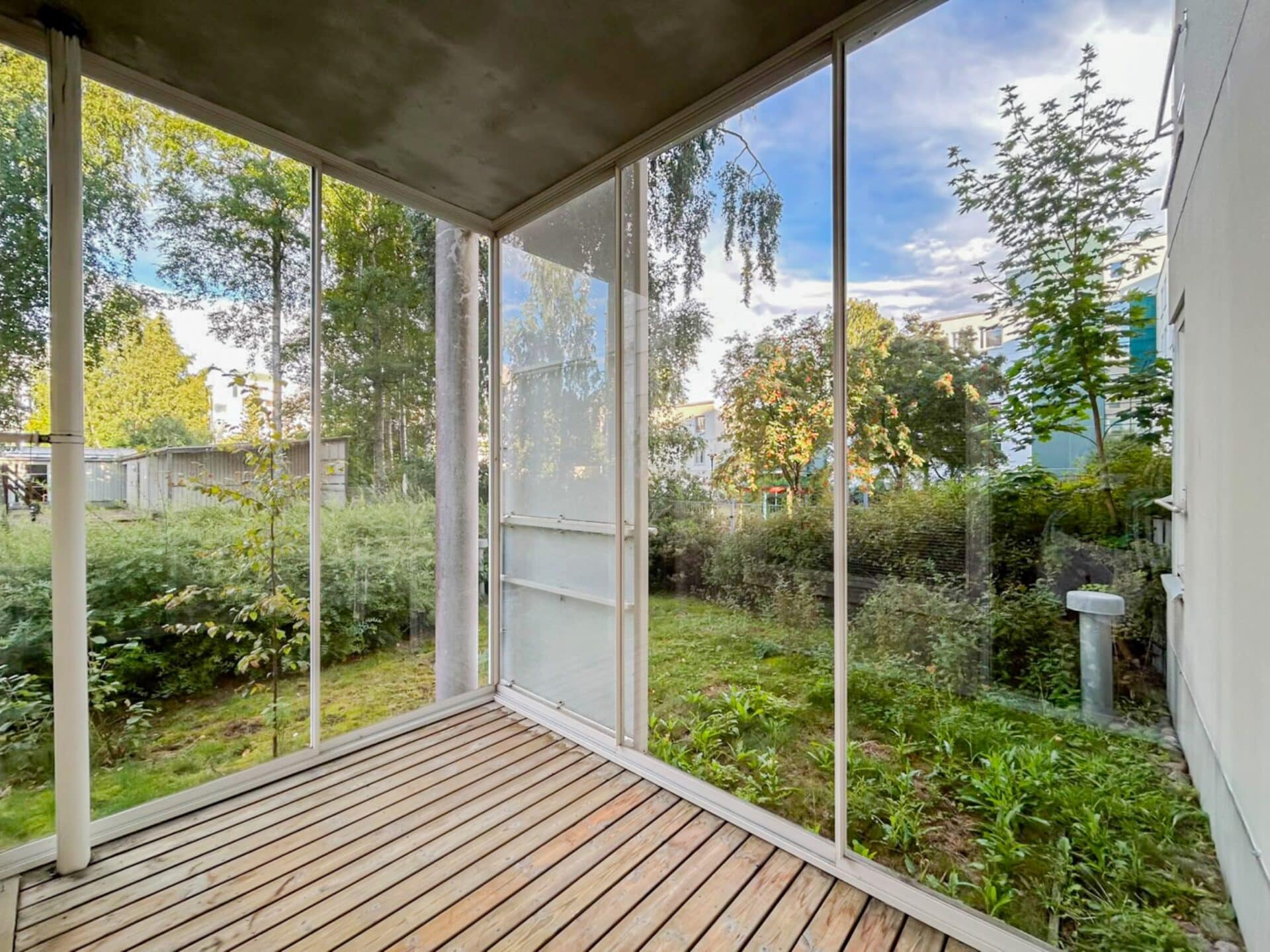 Pieni, katettu puuterassi on lasiseinien ympäröimä, ja sieltä avautuu näkymä vihreälle pihalle, jossa on pensaita ja puita osittain pilvisen taivaan alla.