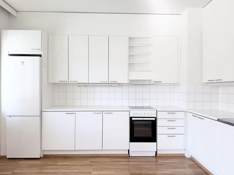 Moderni keittiö, jossa on valkoiset kaapit, valkoinen jääkaappi, uuni ja liesi, laattaseinä sekä puulattia. Tila on siisti, minimalistinen ja valoisa.