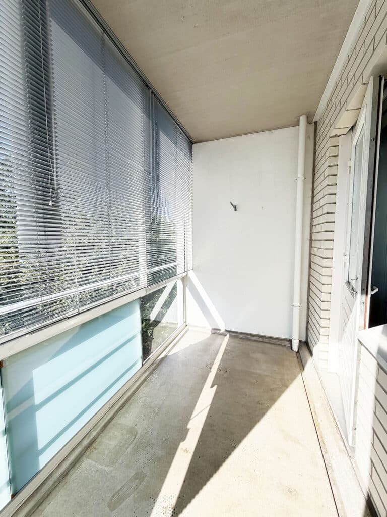 Pieni, tyhjä parveke, jossa on betonilattia; vasemmalla lasi-ikkunat, joiden kaihtimet ovat kiinni, ja oikealla valkoinen tiiliseinä ja ovi. Auringonvalo heijastaa varjoja lattialle.