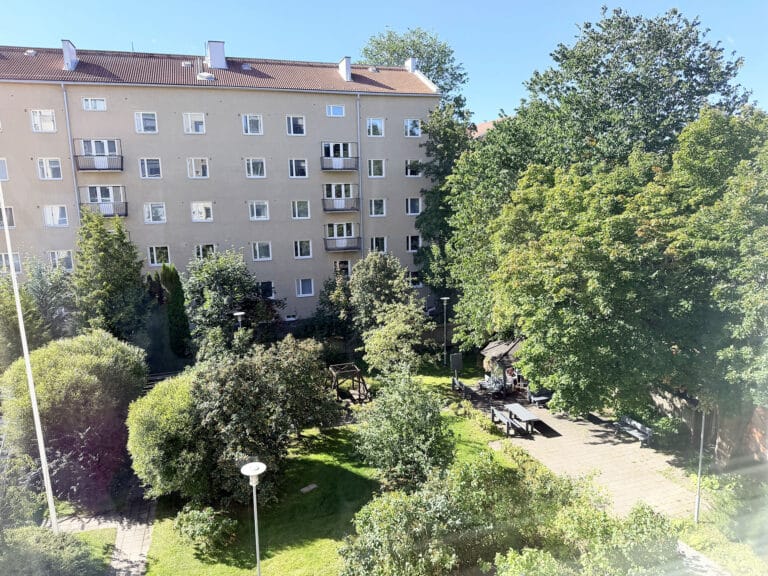Aurinkoinen sisäpiha, jossa on vihreitä puita, penkkejä ja istuvia ihmisiä; taustalla kirkkaan sinisen taivaan alla näkyy beige kerrostalo, jossa on parvekkeita ja paljon ikkunoita.