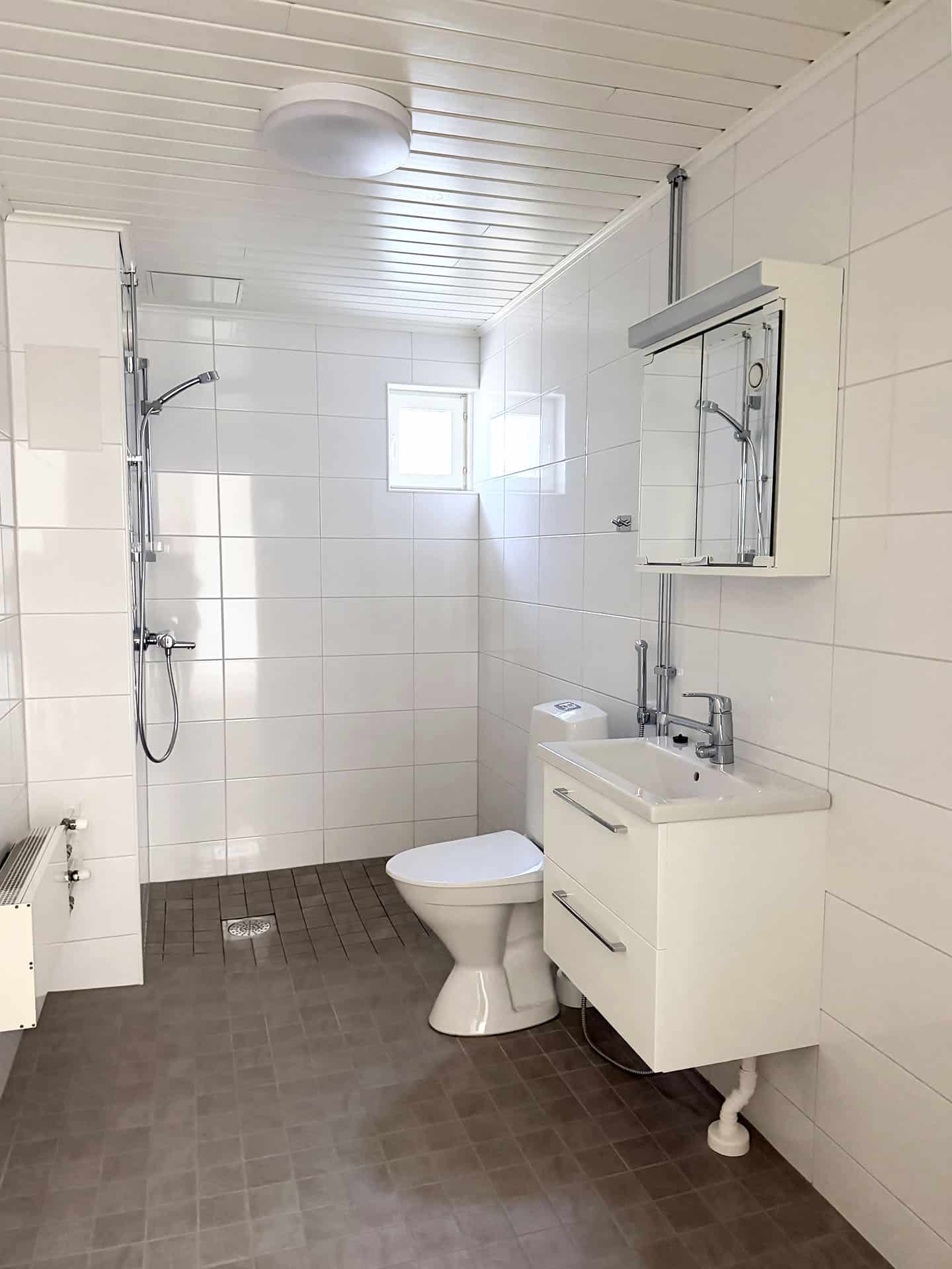 Siisti, moderni kylpyhuone, jossa on valkoisella laatoilla päällystetyt seinät, suihku, wc, valkoinen pesuallaskaappi, peilikaappi ja pieni ikkuna, josta tulvii luonnonvaloa.