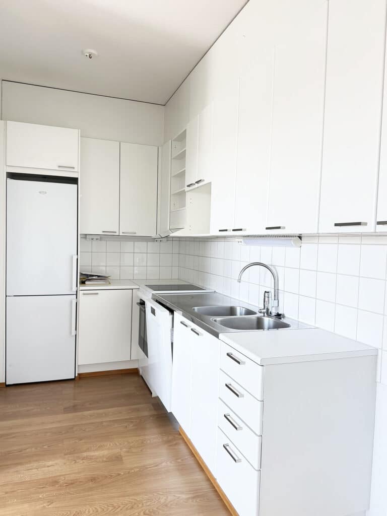 Moderni keittiö, jossa on valkoiset kaapit, kaksialtainen pesuallas, sähköliesi, astianpesukone, jääkaappi ja uuni. Lattia on puuta, ja seinän alaosa on laatoitettu valkoisilla laatoilla. Tila vaikuttaa siistiltä ja valoisalta.
