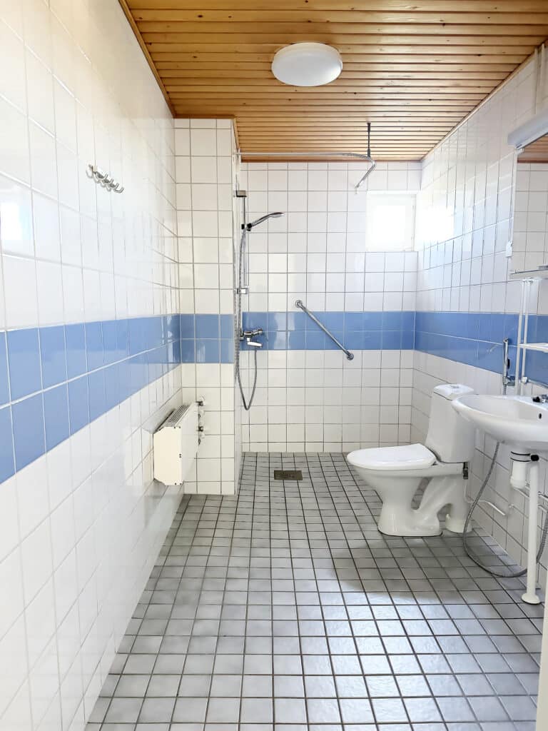Siisti ja esteetön kylpyhuone, jonka seinät on laatoitettu valkoisilla ja sinisillä laatoilla. Siellä on wc, pesuallas, suihku, jossa on tukikaide, seinäpeili ja puinen katto. Luonnonvaloa tulee sisään pienestä ikkunasta.