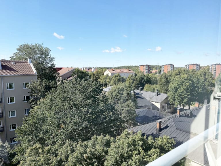 Näkymä korkealta ikkunasta, jossa näkyy kattoja, puita ja kerrostaloja kirkkaan sinisen taivaan alla, jossa on muutamia pilviä.