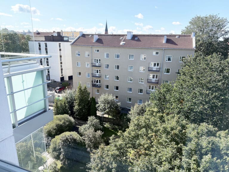 Näkymä korkealta ikkunasta sisäpihalle, jossa on vihreitä puita ja pensaita. Taustalla näkyy beige kerrostalo ja vasemmalla moderni lasirakennus kirkkaan sinisen, pilvisen taivaan alla.