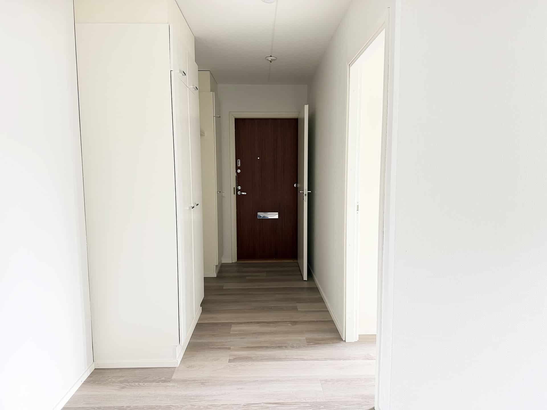 Eteinen, jossa on vaaleat puulattiat, valkoiset seinät, vaatekaapit vasemmalla ja ruskea ovi käytävän päässä. Oikealla on avoin oviaukko. Tila vaikuttaa siistiltä ja minimalistiselta.
