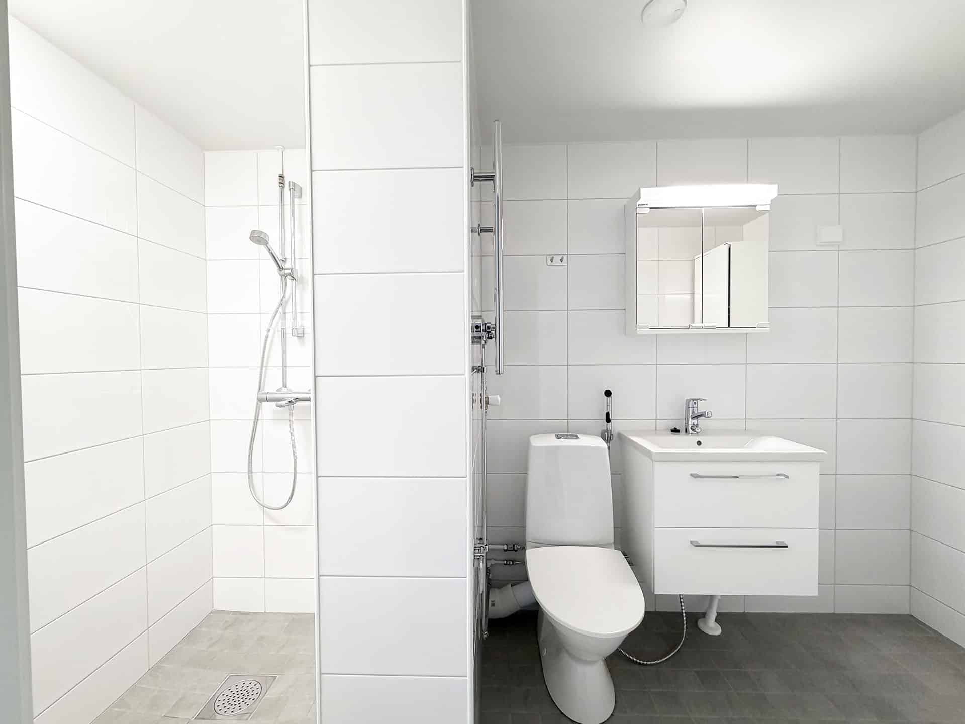 Moderni valkoinen kylpyhuone, jossa vasemmalla on suihkualue, keskellä wc ja oikealla pesuallas, jonka vieressä on kaappi ja peilikaappi. Huoneen seinät on laatoitettu ja lattia on harmaa.