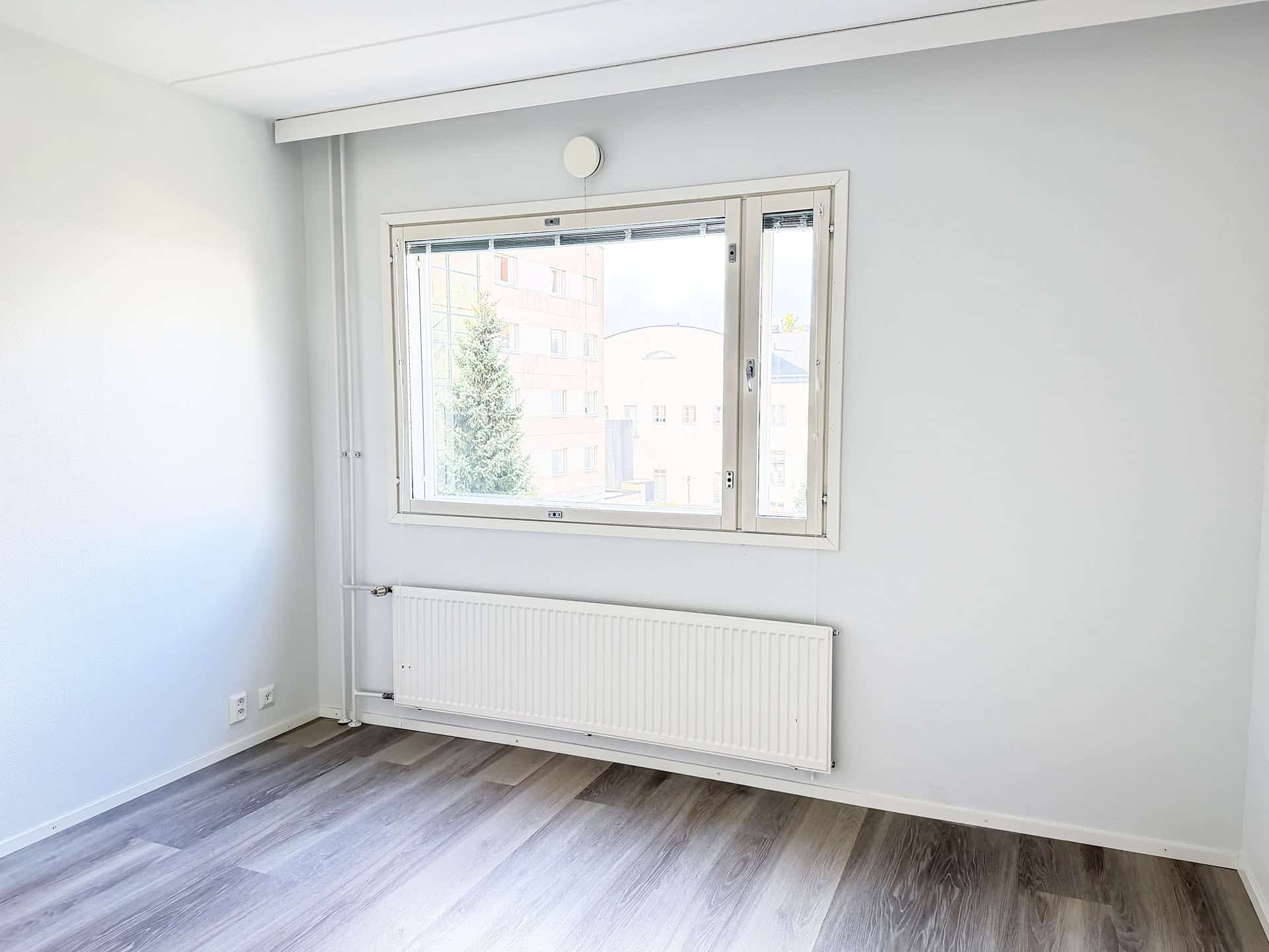 Valoisa, tyhjä huone, jossa on vaaleanharmaat seinät, puukuvioinen lattia, suuri ikkuna, josta avautuu näkymä rakennuksiin ja puihin, sekä valkoinen lämpöpatteri ikkunan alla.