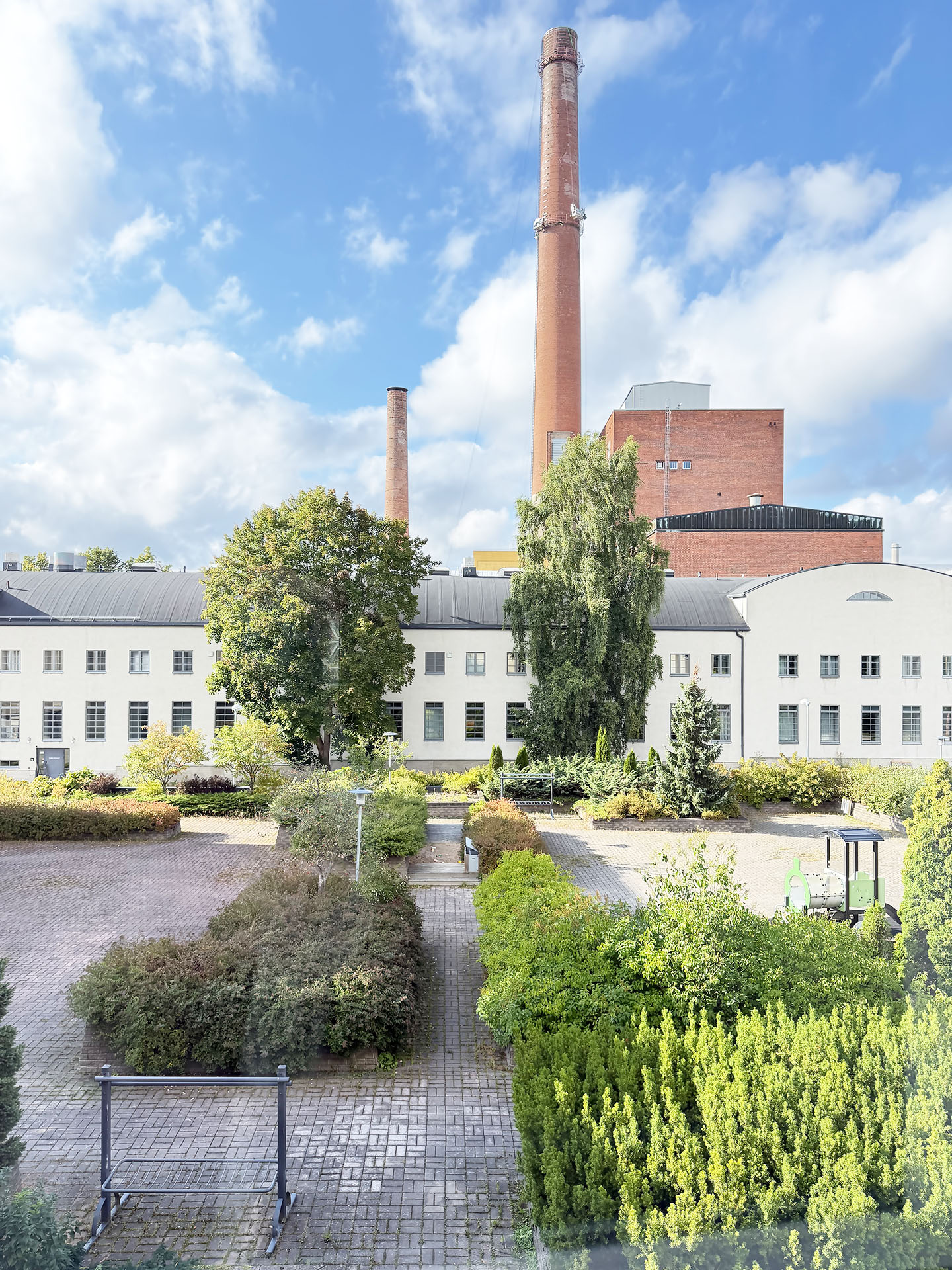 Ulkona on kirkas, aurinkoinen päivä, ja edessä kohoaa suuri valkoinen teollisuusrakennus, jossa on korkeat tiiliset savupiiput; rakennusta ympäröivät vihreät puut, pensaat ja kivetty piha.