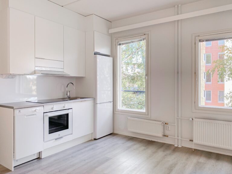 Minimalistinen valkoinen keittiö, jossa on sisäänrakennettu uuni, liesi, jääkaappi sekä kaksi ikkunaa, joista avautuu näkymä puille ja viereiselle rakennukselle.