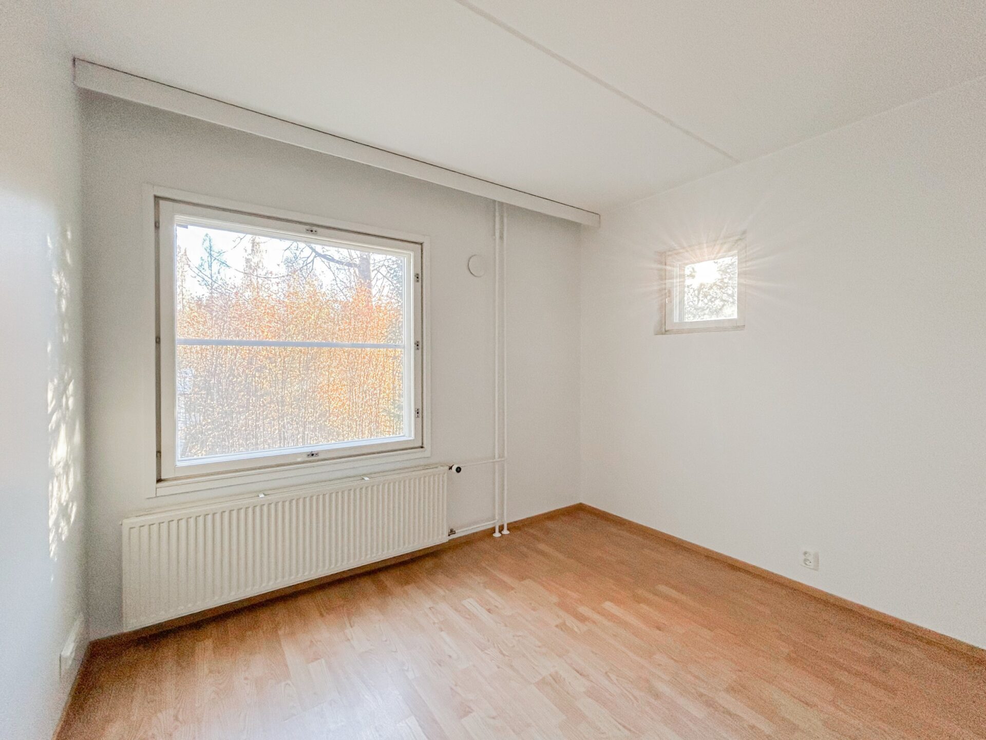 Tyhjä huone, jossa on valkoiset seinät, puulattia, suuri ikkuna, josta avautuu näkymä puille, sekä pienempi neliönmuotoinen ikkuna.