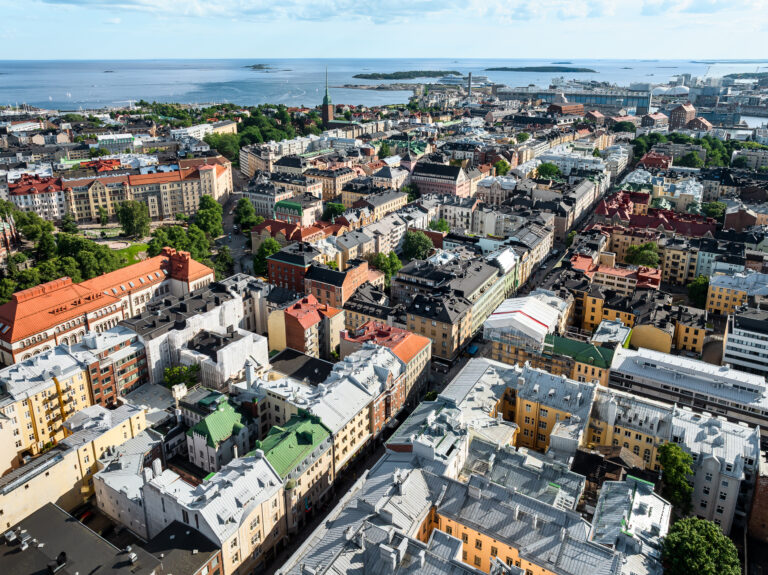 Ilmakuva tiheästi rakennetusta eurooppalaisesta kaupungista, jossa on monikerroksisia rakennuksia sekä punaisia ja harmaita kattoja, ja jonka taustalla näkyy satama ja meri.