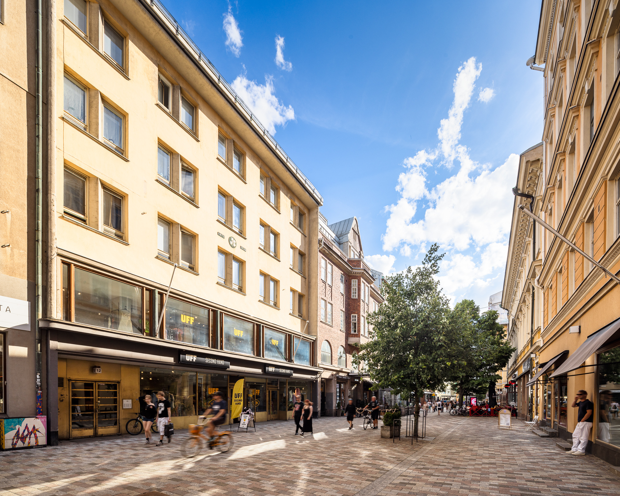 Aurinkoinen, kävelykatu, jota reunustavat monikerroksiset rakennukset ja liiketilat, mukaan lukien UFF-kirpputori, ja jota pitkin useat ihmiset kävelevät ja pyöräilevät kivettyä polkua pitkin.