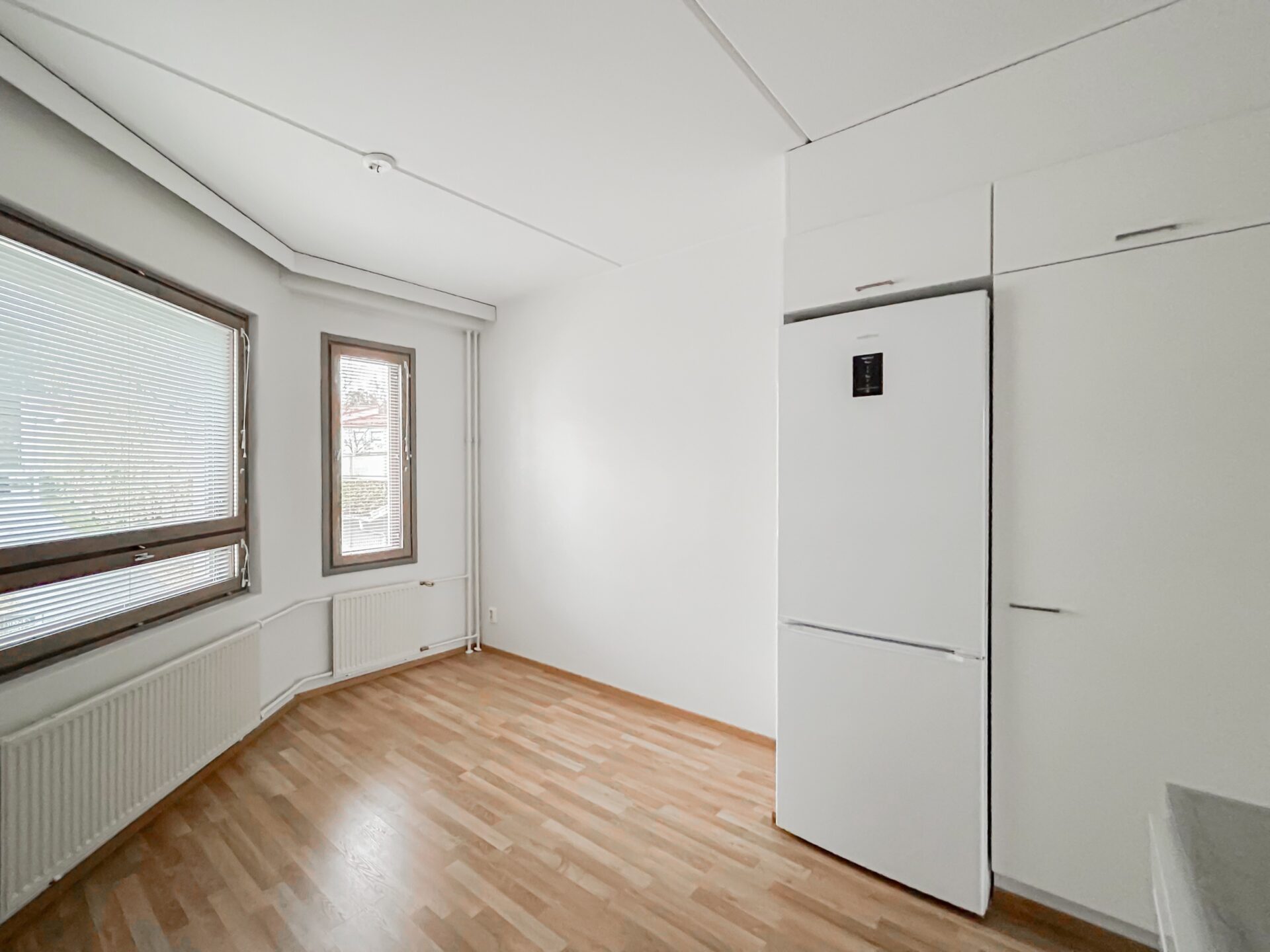 Minimalistinen huone, jossa on puun näköinen lattia, kaksi ikkunaa kaihtimilla, valkoinen lämpöpatteri sekä kaapistoon integroitu korkea valkoinen jääkaappi.
