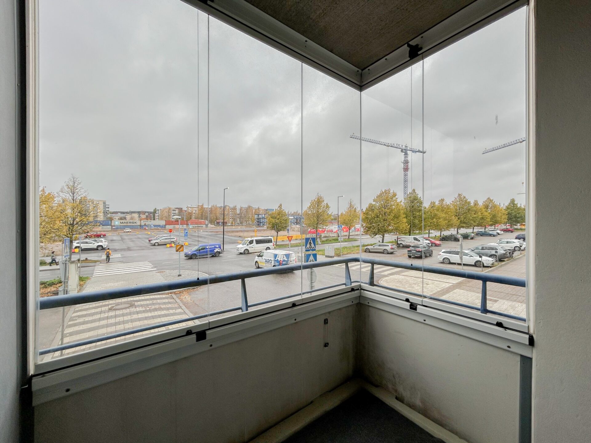 Näkymä lasitettuun parvekkeelta, josta avautuu näkymä katujen risteykseen, pysäköintialueelle ja rakennustyömaalle pilvisen taivaan alla.