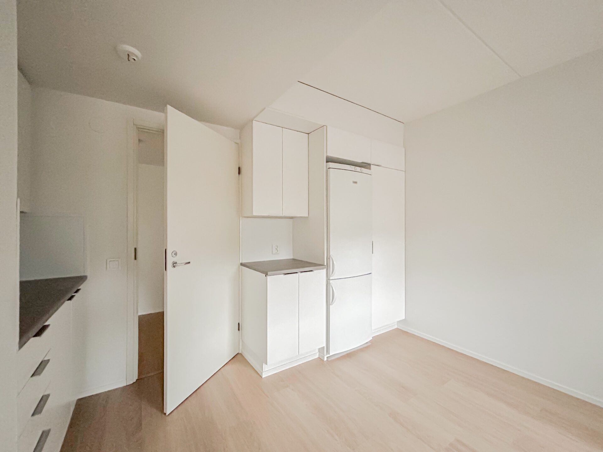 Minimalistinen keittiö, jossa on valkoiset kaapit, jääkaappi ja avoin oviaukko, joka johtaa toiseen huoneeseen.