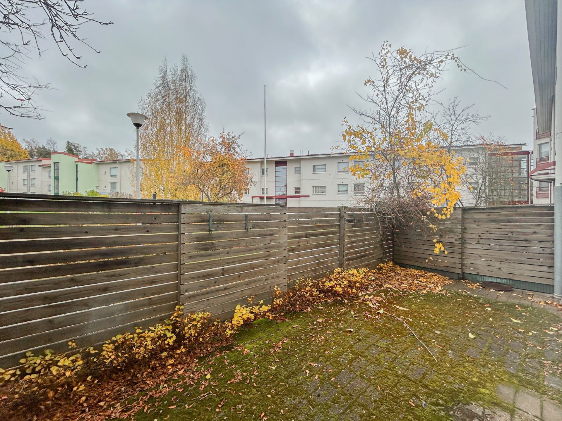 Aidattu terassialue, jolla on hajallaan syksyn lehtiä, ja taustalla näkyvä kerrostalo pilvisen taivaan alla.