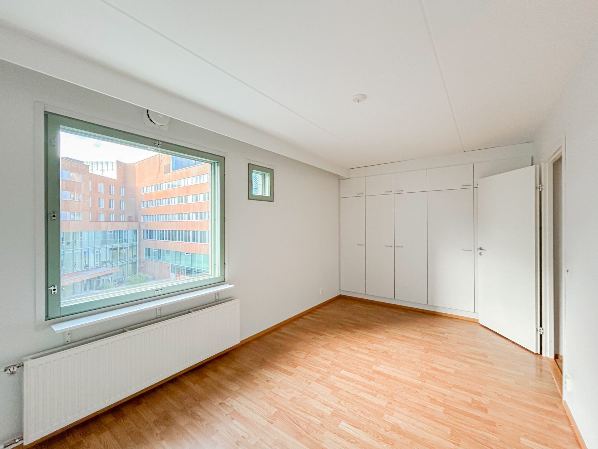 Tyhjä huone, jossa on puun näköinen lattia, suuri ikkuna, josta avautuu näkymä moderniin rakennukseen, sekä seinä täynnä valkoisia kiinteitä kaappeja.