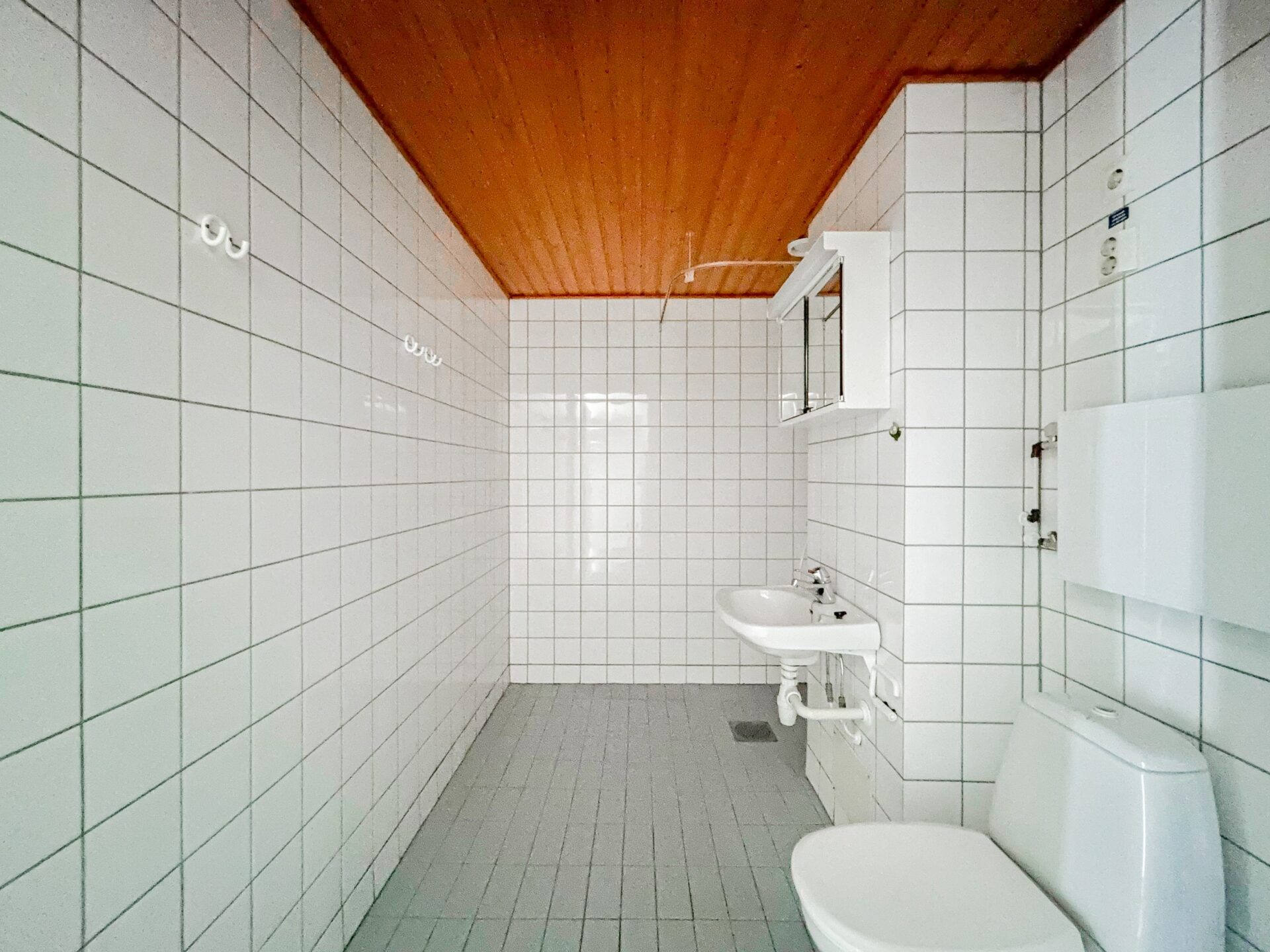 Pieni kylpyhuone, jossa on valkoisella laatoitetut seinät, puinen katto, valkoinen pesuallas ja wc.